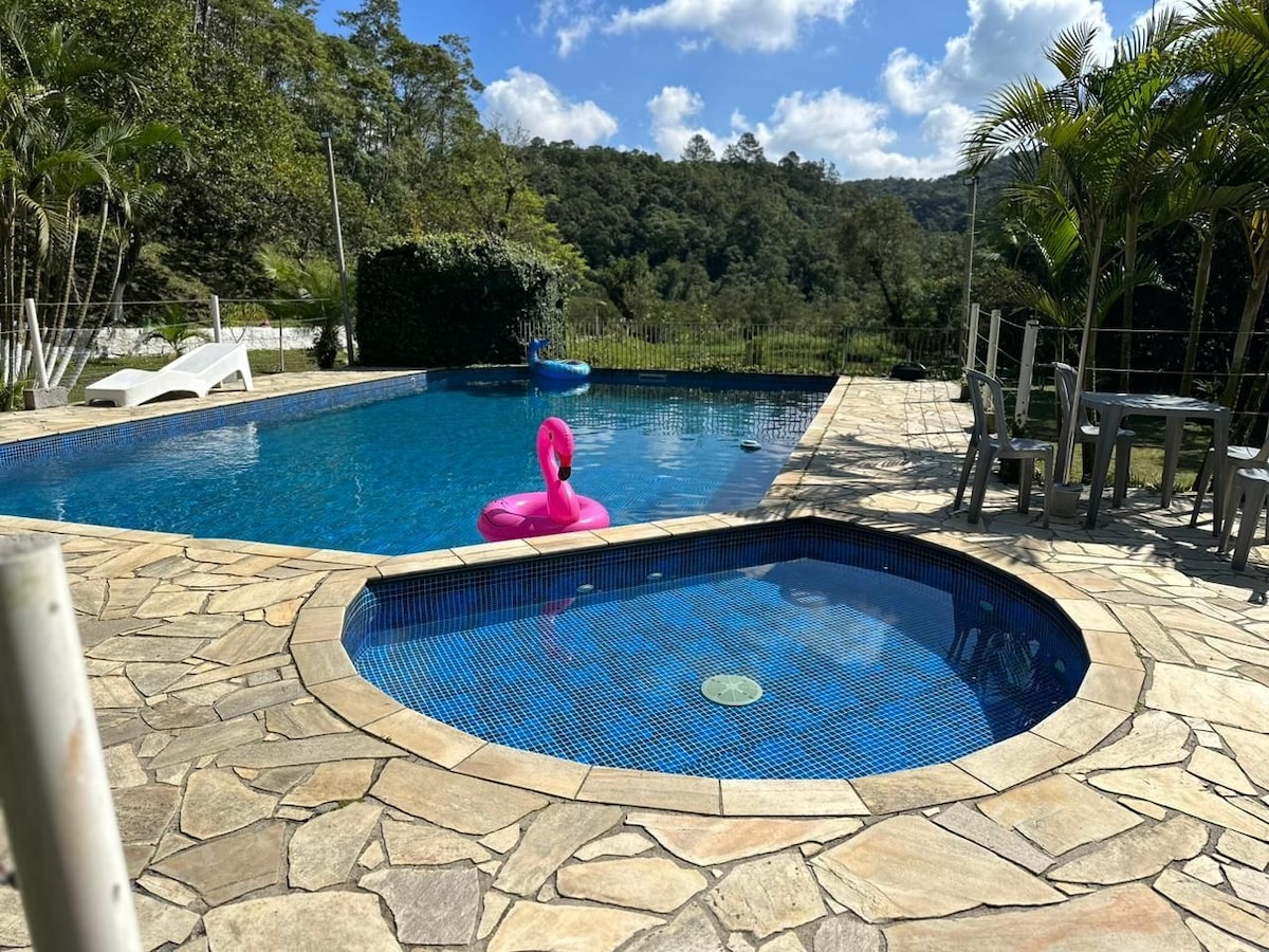 A well-maintained swimming pool area is shown, featuring a large blue pool and a smaller jacuzzi with a tiled edge. Bright umbrellas provide shade around the pool. Lush greenery accompanies the area, enhancing the natural setting.