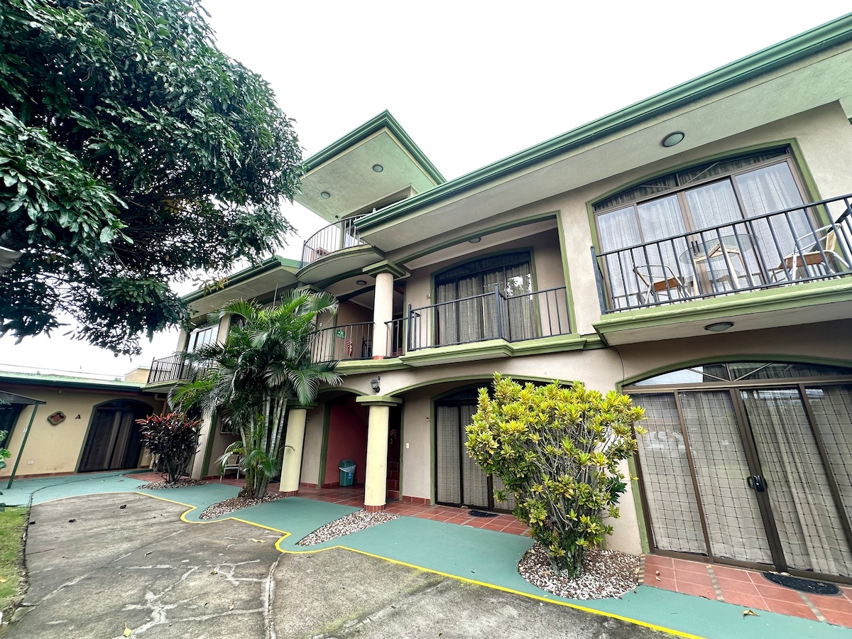 A three-story building with a green roof and multiple balconies is surrounded by lush greenery. Paved walkways with decorative stones connect different areas, while large windows invite natural light indoors, suggesting a welcoming atmosphere for guests.