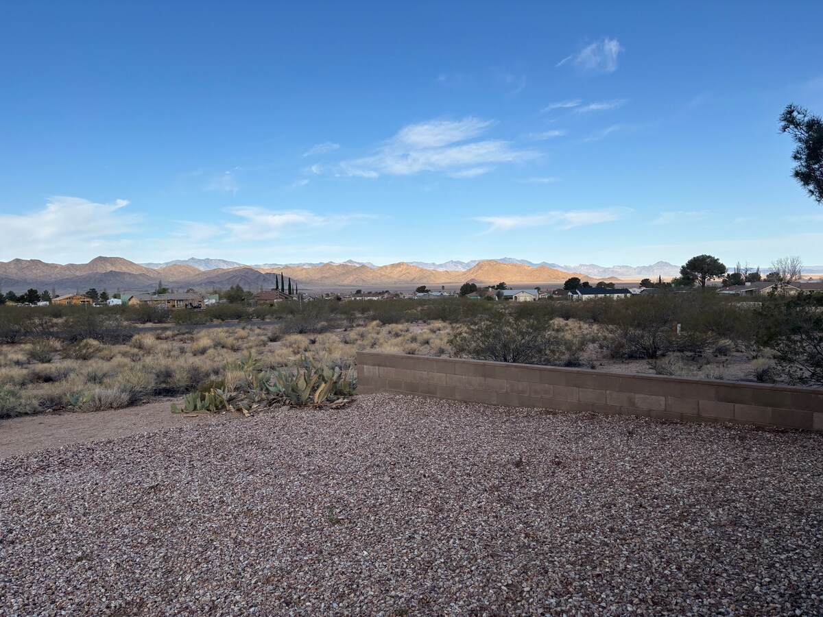 A wide view of the surrounding landscape features gentle elevations and vast vegetation. A rocky, gravelly area is visible in the foreground, leading toward distant mountains under a clear blue sky, offering a sense of openness and tranquility.