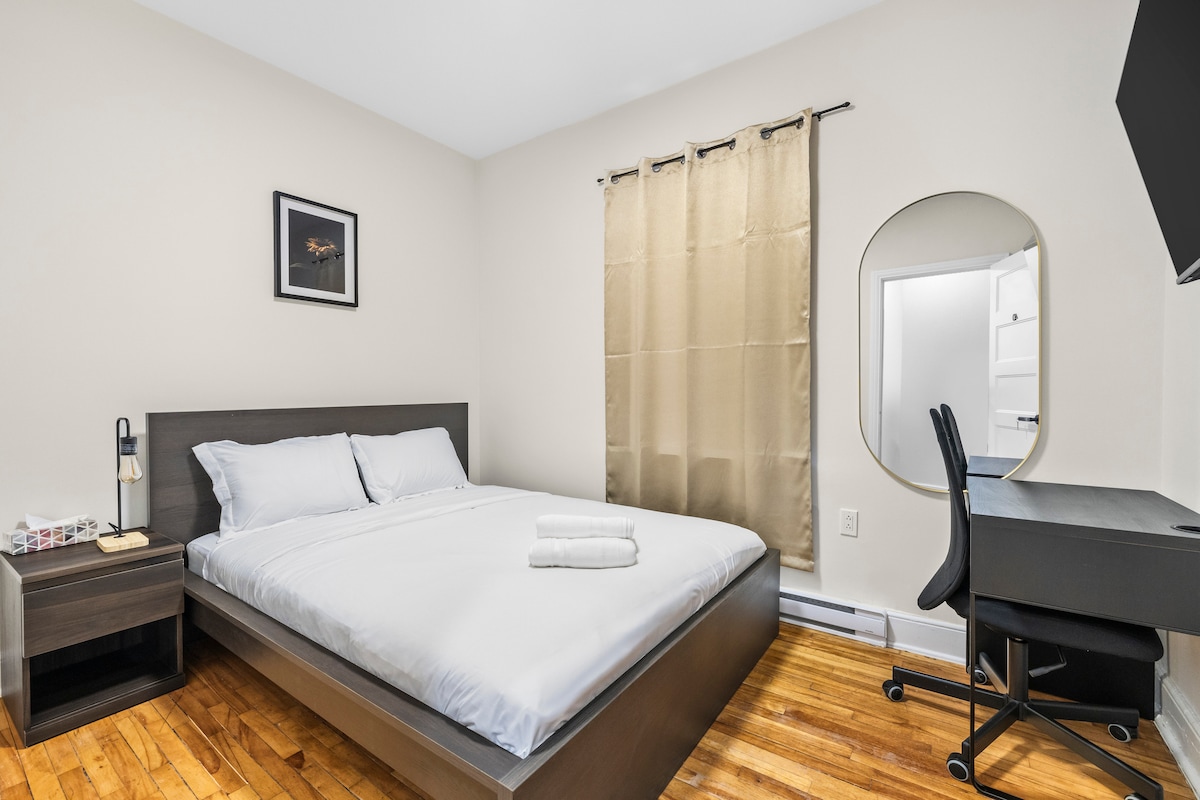 A cozy bedroom features a spacious bed with crisp white linens and neatly arranged towels. A wooden desk with a chair is positioned to the side, while a round mirror reflects the light from the window, which is dressed with beige curtains.