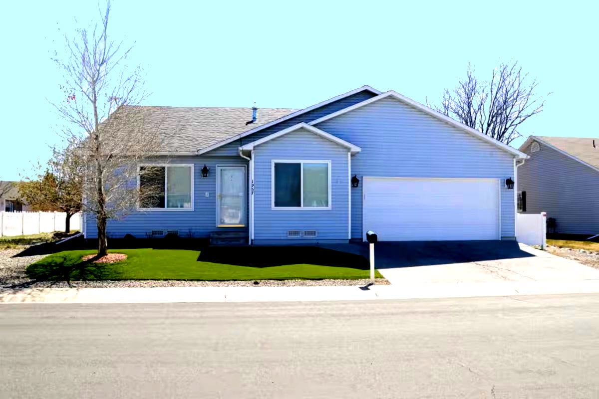 A single-story house features light blue siding and a well-maintained front lawn. The entryway is framed by two medium-sized windows, and a driveway leads to a garage. A mailbox stands at the curb, completing the inviting front view of the home.
