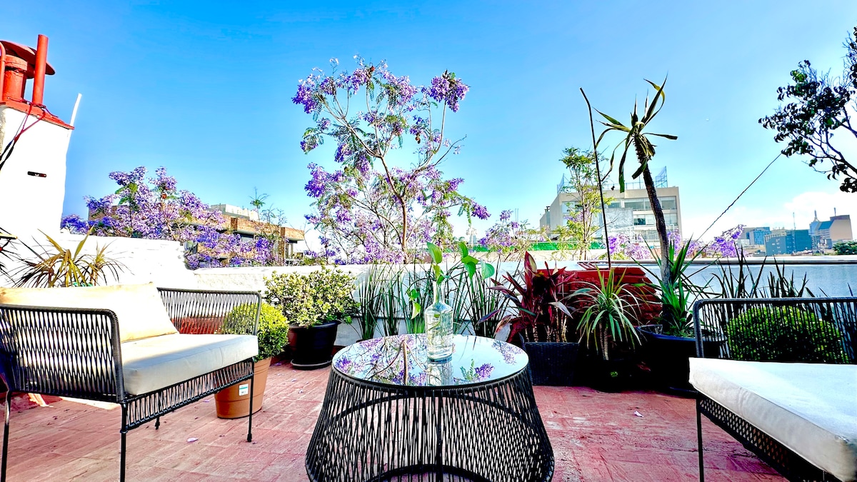 A spacious terrace features a circular black table surrounded by two light-colored seating areas. Lush greenery and a blooming Jacaranda tree provide vibrant purple accents against a clear blue sky. The outdoor space invites relaxation and enjoyment of the surrounding garden-like atmosphere.