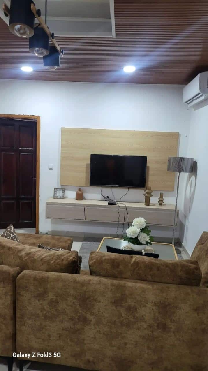 A comfortable living area is designed with a plush brown couch and a coffee table adorned with a decorative arrangement of flowers. A wall-mounted television is visible above a light wood media unit, complemented by ambient lighting and a stylish lamp.