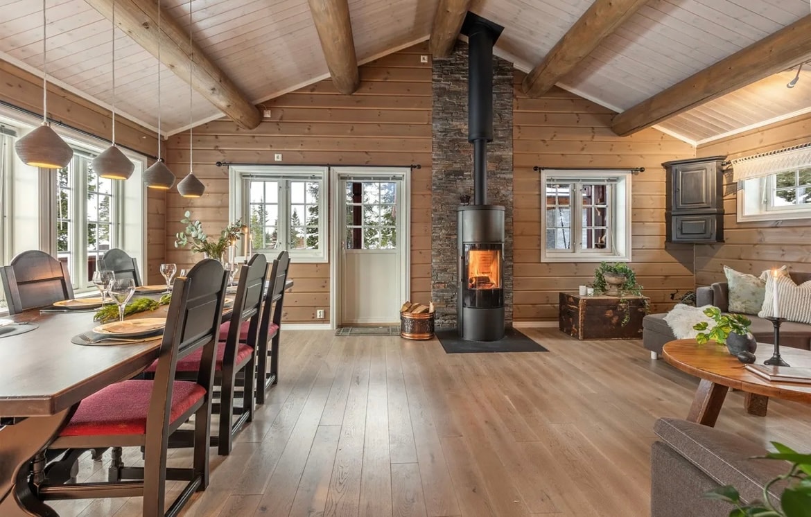A spacious living area features warm wooden walls and a high ceiling supported by wooden beams. A dining table accommodates eight chairs, and a cozy seating area is located near a stone fireplace, providing a central point for gatherings. Large windows allow natural light and views of the outdoors.