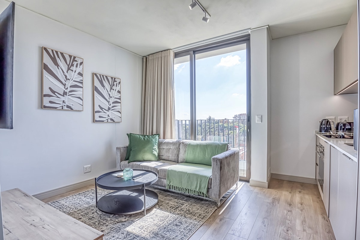 A bright living area features a comfortable sofa with a mix of green and grey upholstery. Two framed botanical prints adorn the wall. Natural light fills the space through large glass doors, offering a view of the outdoors. A round coffee table sits on a textured rug.