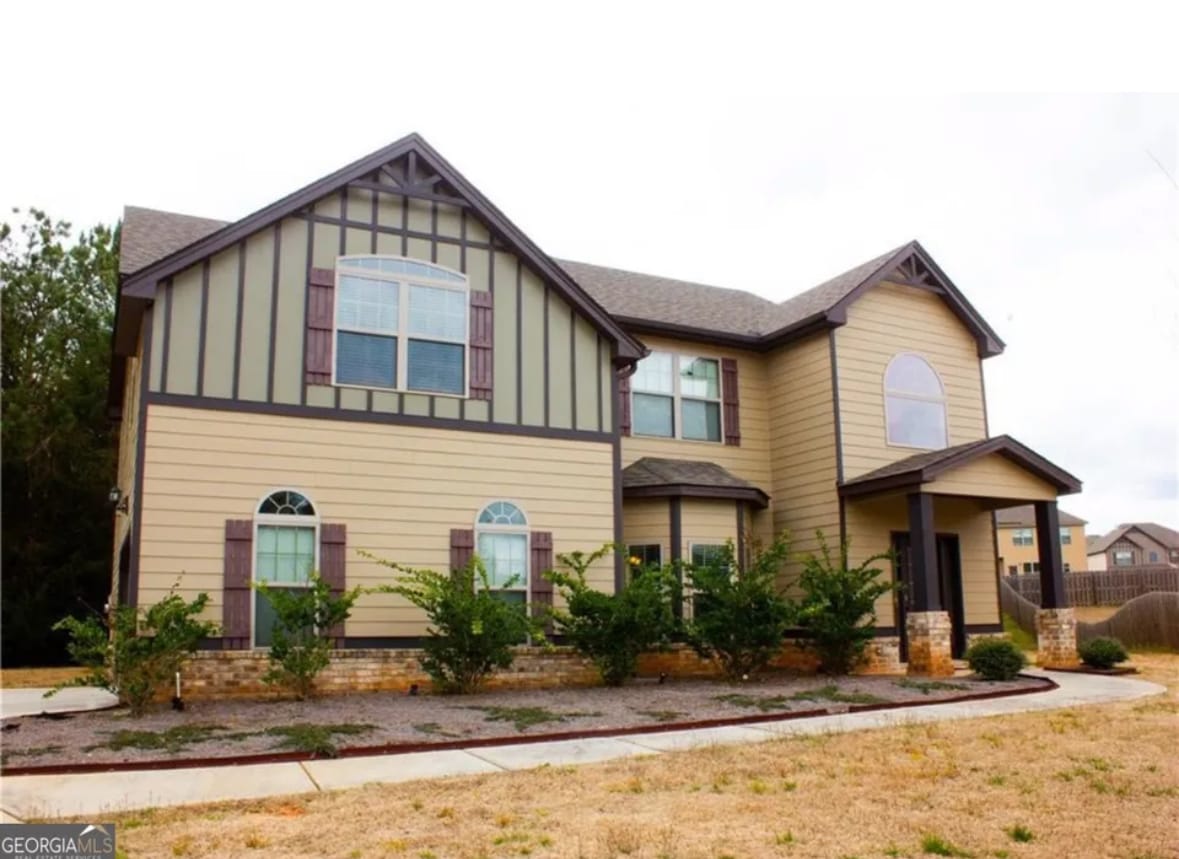 The exterior of the house features a blend of light brown and cream siding, accented by decorative wooden shutters. Symmetrical windows offer a balanced appearance, while a welcoming entrance is framed by a small porch. Landscaped shrubbery enhances the front yard view.