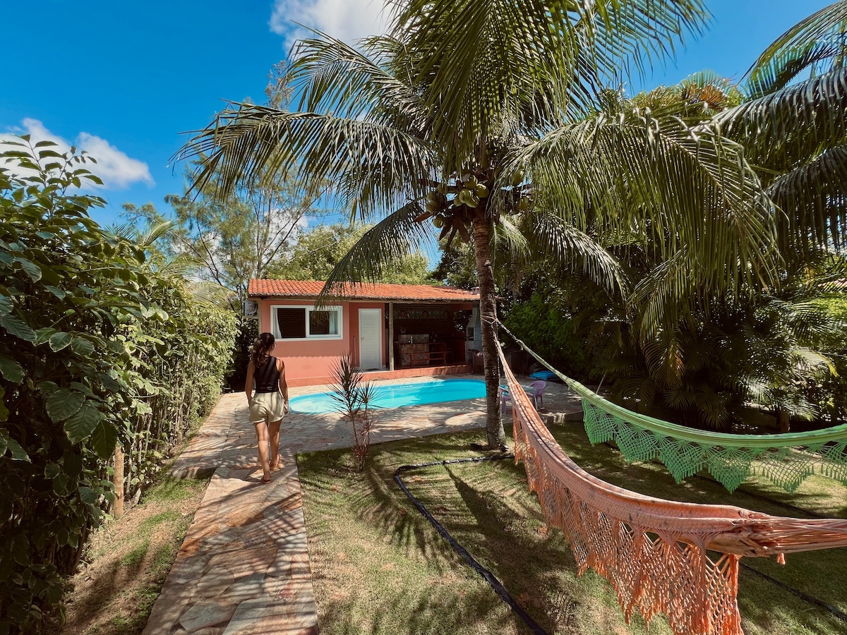 A peaceful outdoor space is surrounded by lush greenery, featuring a clear swimming pool and a hammock. A stone pathway leads toward a cozy flat, which is partially visible amidst palm trees under a bright blue sky.