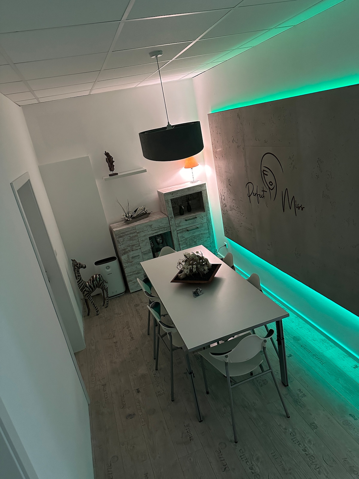 A modern communal space is presented, featuring a long white dining table accompanied by several chairs. A softly lit pendant light hangs above. Ambient green lighting outlines the wall, where a logo is displayed. The floor is covered in wooden planks, adding warmth to the area.
