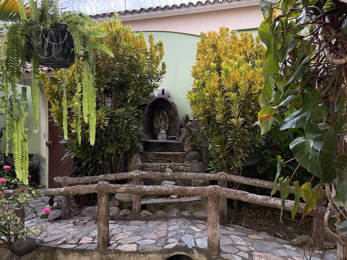 A serene garden area is visible, featuring a pathway of stone leading to a small water feature and statue. Lush greenery surrounds the path, with various plants and flowers enhancing the tranquil atmosphere. A rustic wooden bridge crosses over the pathway, blending with the natural setting.