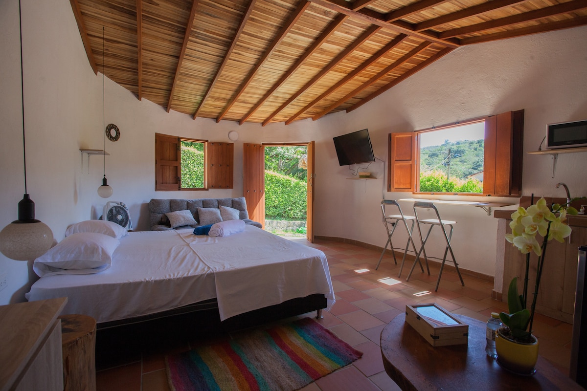 A cozy interior space features a spacious bed with soft linens, accompanied by decorative pillows. Natural light streams through two open windows, illuminating the earthy tones of the room. A small kitchen area is equipped with essential appliances, and a wooden table with two chairs provides a casual seating option.