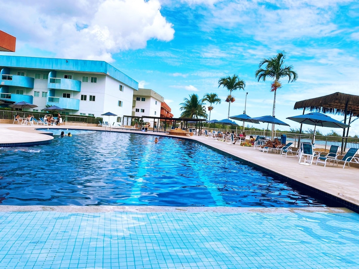 A large swimming pool is depicted, featuring clear blue water surrounded by lounge chairs and umbrellas. Tropical palm trees are visible in the background. Several guests are enjoying the pool area under a sunny sky with scattered clouds.