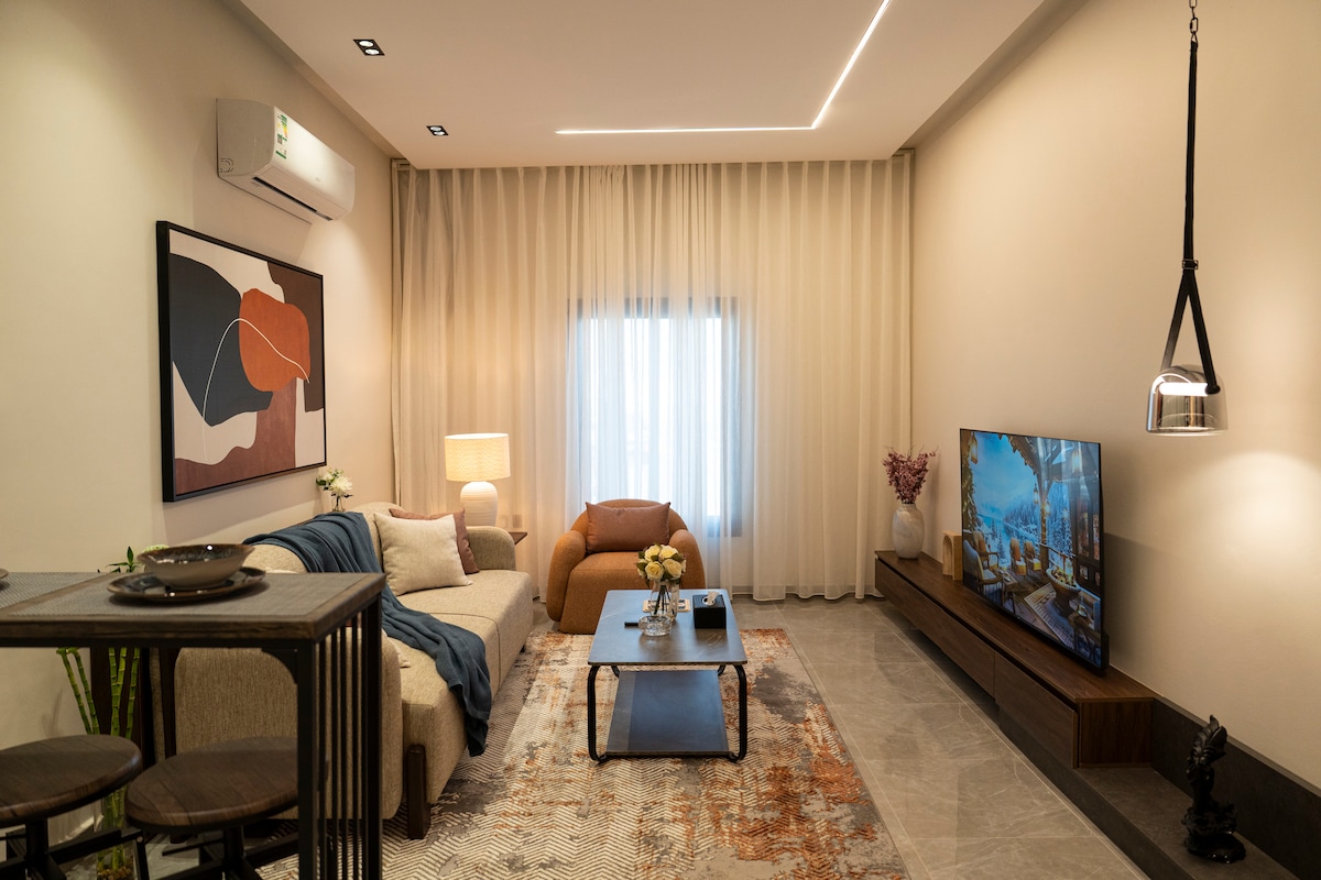The living area features a soft beige sectional sofa paired with an orange accent chair. A coffee table sits in the center on a patterned rug. A smart TV is mounted on the wall opposite a decorative console, while soft drapes frame the window allowing natural light to enter.