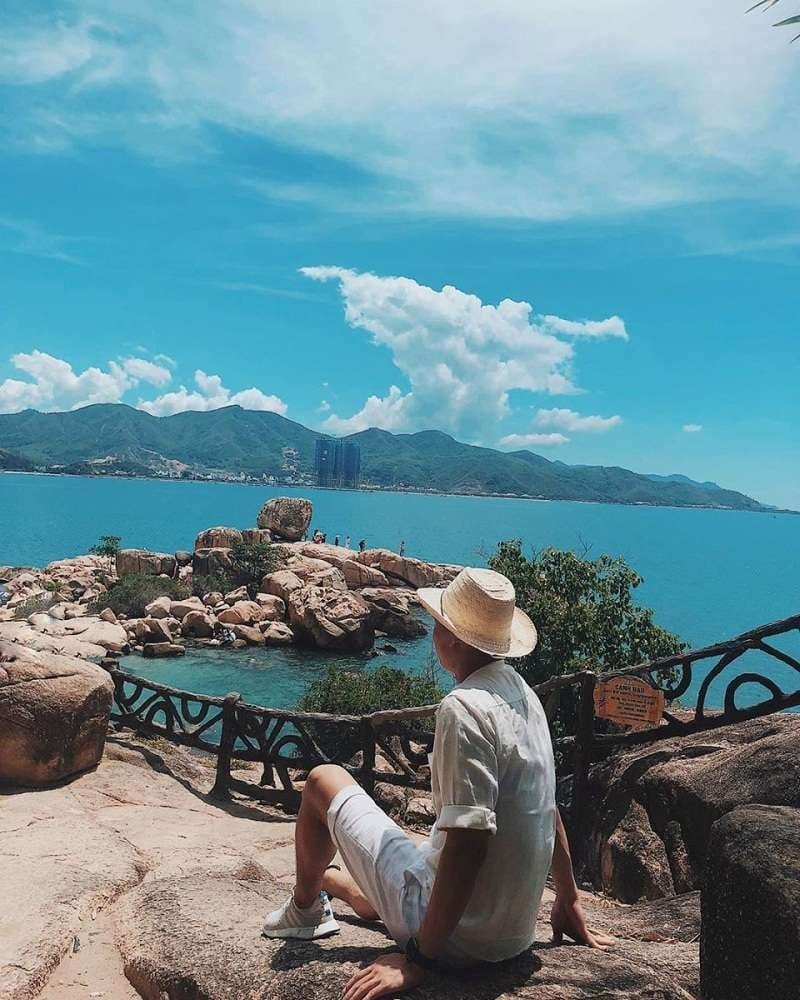 A person is seated on a rocky ledge, overlooking a calm blue sea dotted with stones. Lush green hills rise in the background against a bright blue sky marked with fluffy white clouds. A winding black railing frames the scene, enhancing the natural beauty.