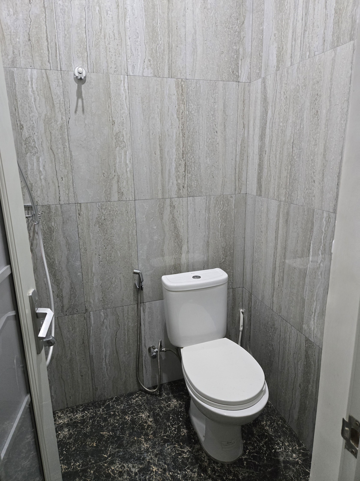 A modern bathroom is featured with polished gray stone walls and a dark patterned floor. A white toilet is positioned against the wall, complemented by a handheld shower attachment. A small hook is mounted on the wall for convenience.