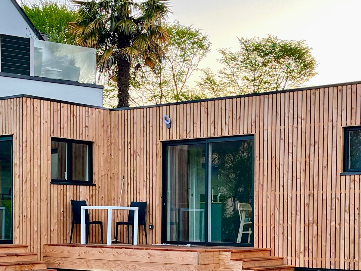A wooden exterior is presented, characterized by vertical slats that provide a natural appearance. Two outdoor chairs and a small table are arranged on a wooden deck, inviting alfresco dining. Expansive glass doors allow visibility into a modern interior, framed by greenery.