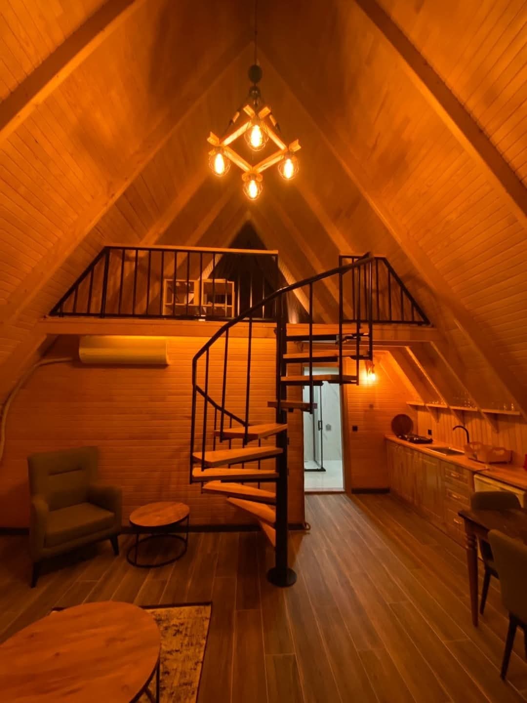 An inviting interior displays vaulted wooden ceilings and warm lighting. A spiral staircase leads to a loft area, while a comfortable seating arrangement comprises a green chair and a small wooden table. The kitchen area features a countertop and modern appliances, enhancing functionality.