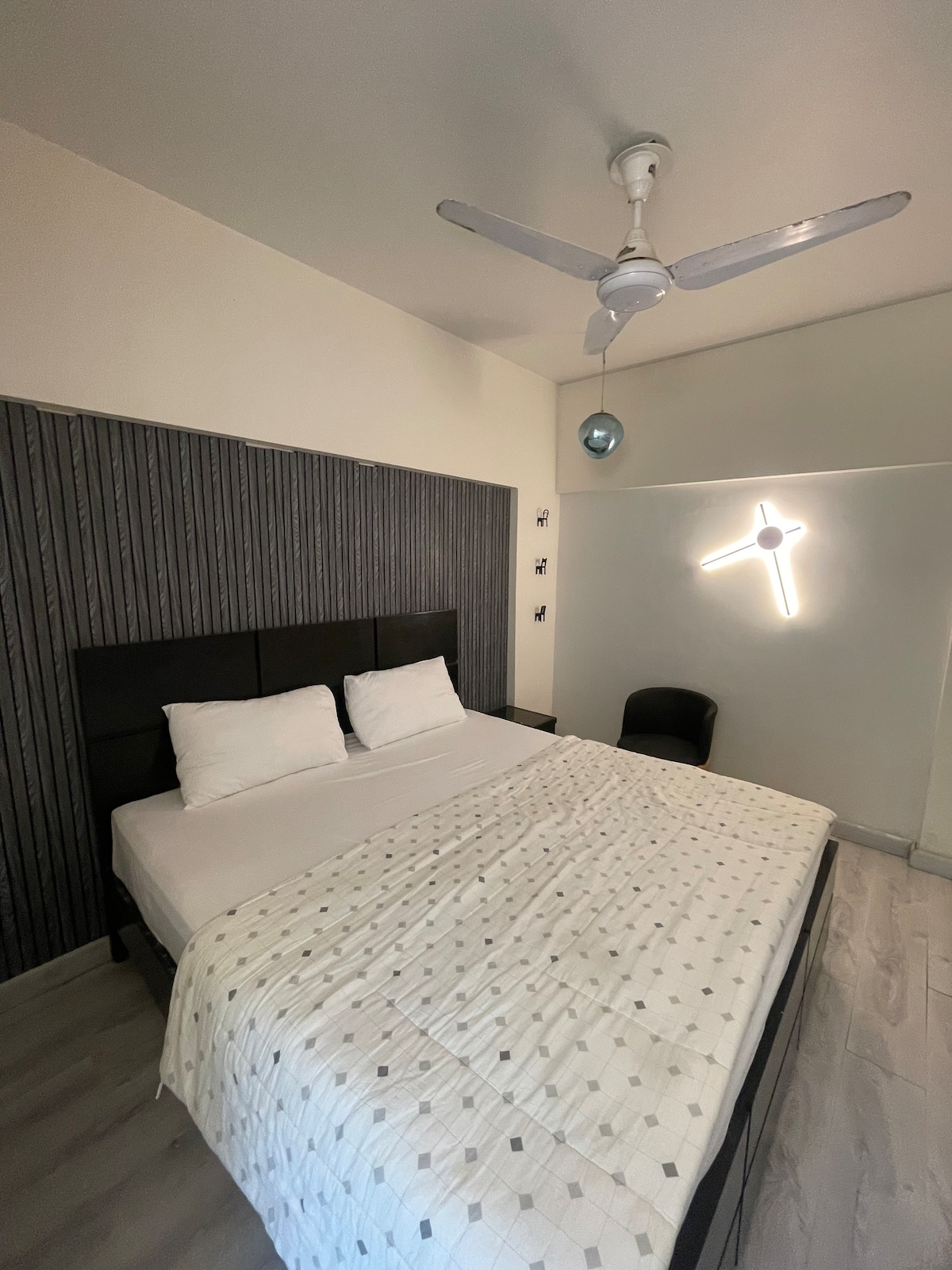 A spacious bedroom features a large bed with white linens and two decorative pillows. The light neutral tones of the walls are complemented by a textured accent wall. A ceiling fan and modern light fixture provide illumination, while a chair is situated in one corner of the room.