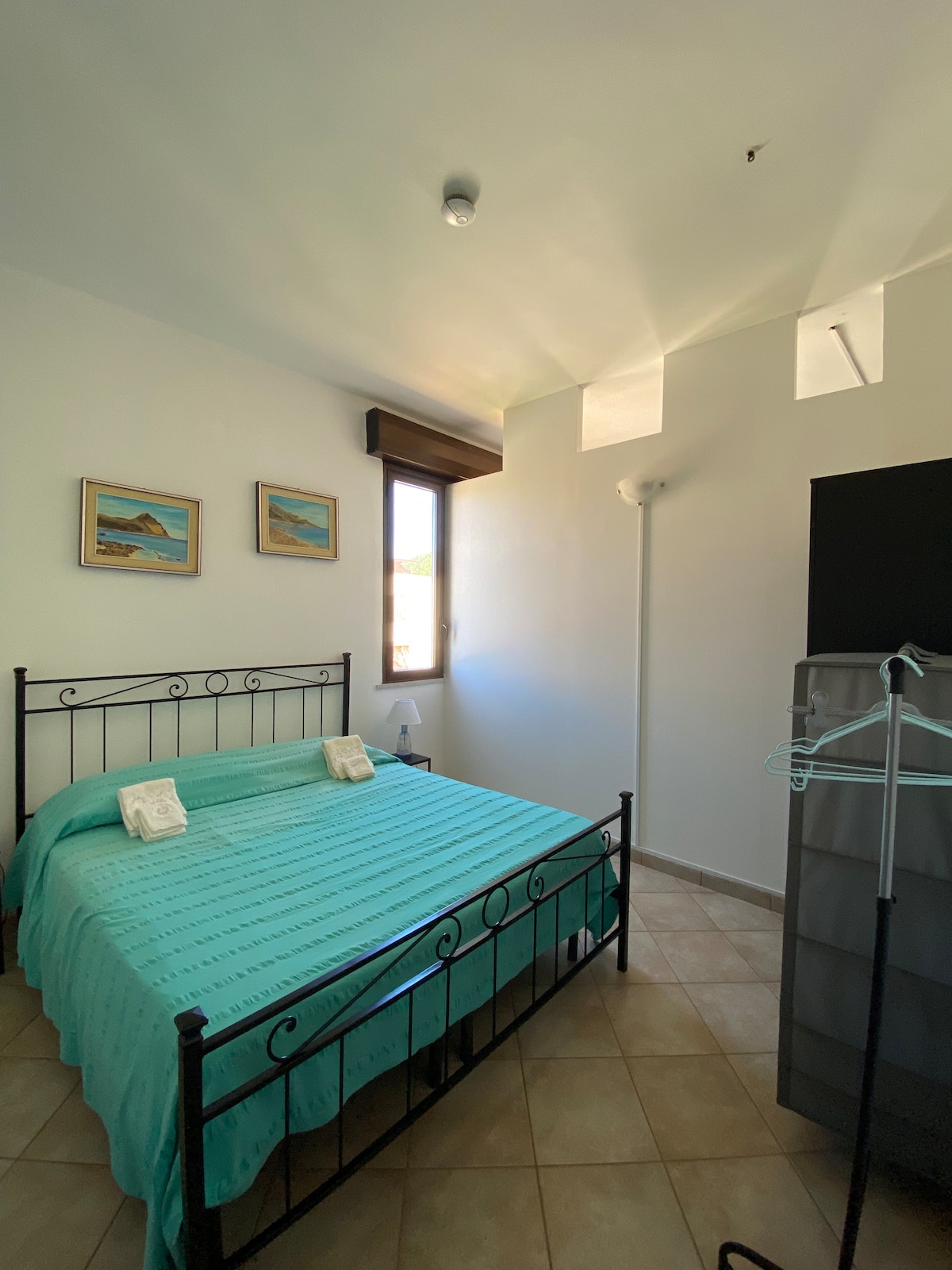A well-lit bedroom features a spacious bed covered in a turquoise quilt. Two framed coastal paintings adorn the white walls. Natural light streams in through a window, and a tall dark wardrobe is positioned nearby, along with a clothing rack.