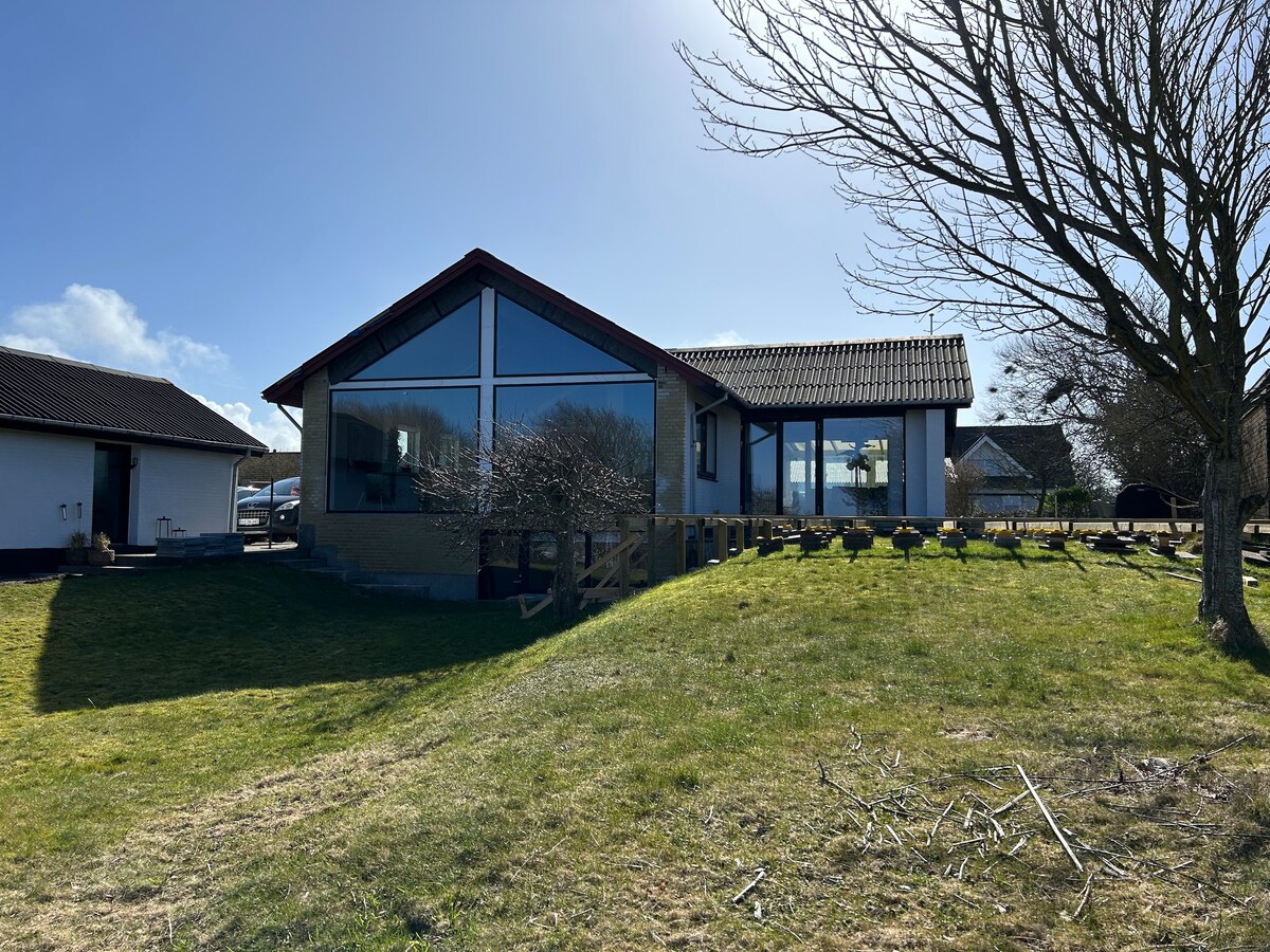 A modern villa is shown with large glass windows reflecting natural light. The exterior features a mix of stone and wood materials. The surrounding grass area is complemented by neatly arranged planters, while a clear blue sky enhances the setting.