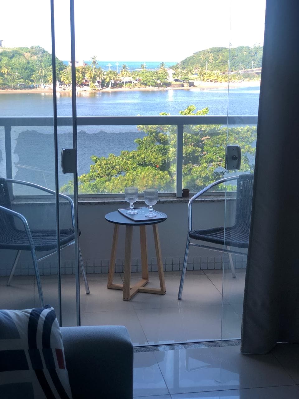 A small balcony area features two gray chairs positioned around a round wooden table. Two clear glasses are placed on the table, with a view of a scenic river and greenery visible through the glass sliding door.