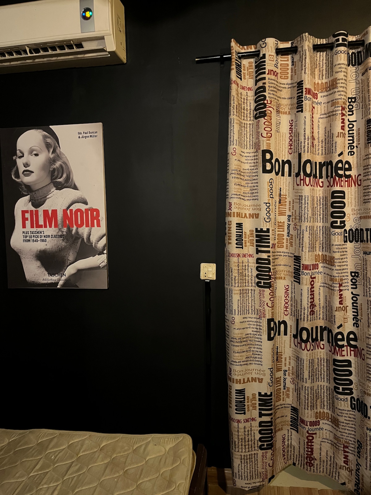 A cozy room features a dark wall, complemented by a poster displaying a film noir theme. A patterned curtain with newspaper-like text covers a window. An air conditioning unit is mounted on the wall, while a spacious bed is positioned nearby.