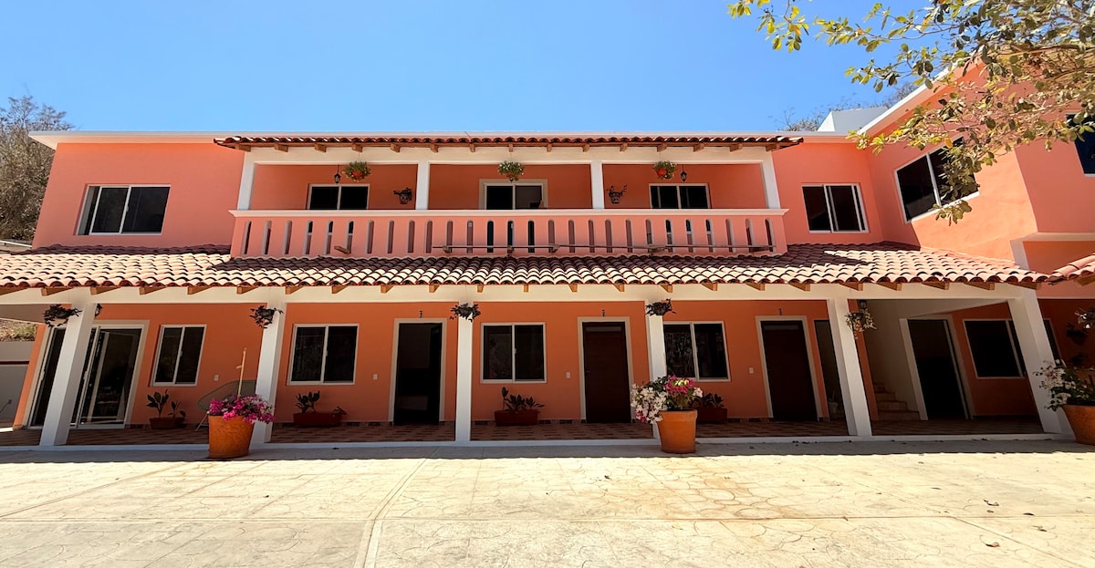 A three-story building with a warm color exterior is seen under a clear blue sky. Potted plants decorate the entrance, while balconies with railings provide an inviting touch. Large windows allow ample natural light to brighten the outdoor space.