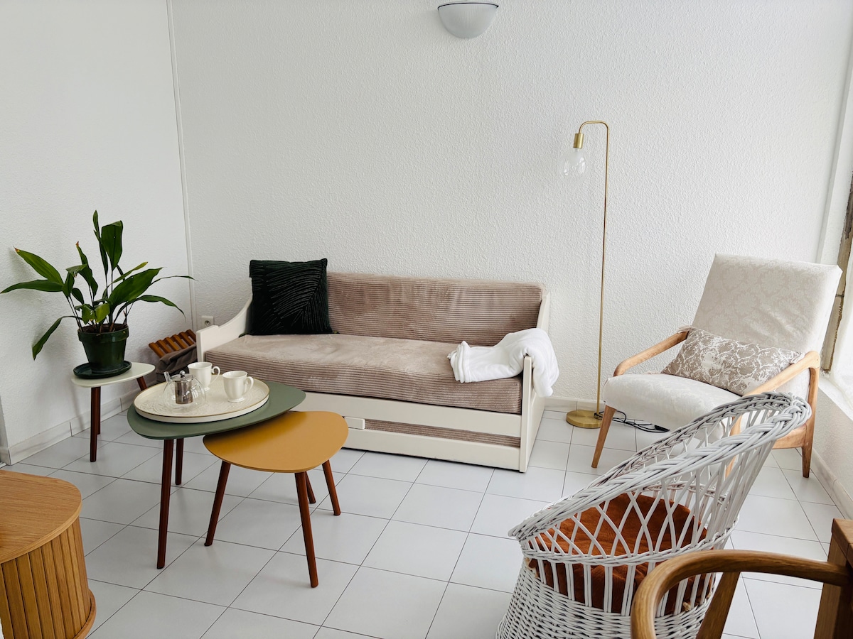 A light-filled living area features a beige sofa and two small side tables in varying shapes. A comfortable armchair is present alongside a wicker chair. A plant adds a touch of greenery, while a floor lamp provides additional lighting.
