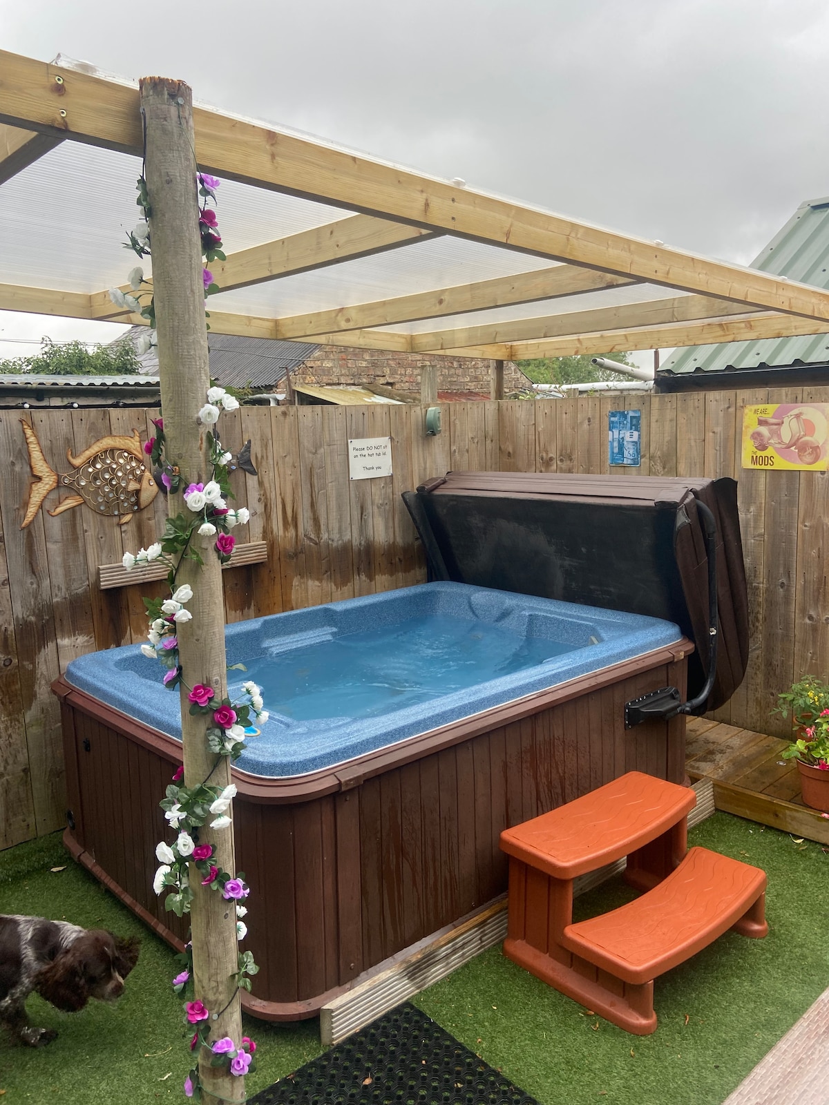 A covered hot tub is positioned within a fenced area, featuring wooden steps for easy access. Colorful flowers are seen climbing along the support posts, and planters add a touch of greenery. The surroundings include artificial grass, providing a clean and inviting space.