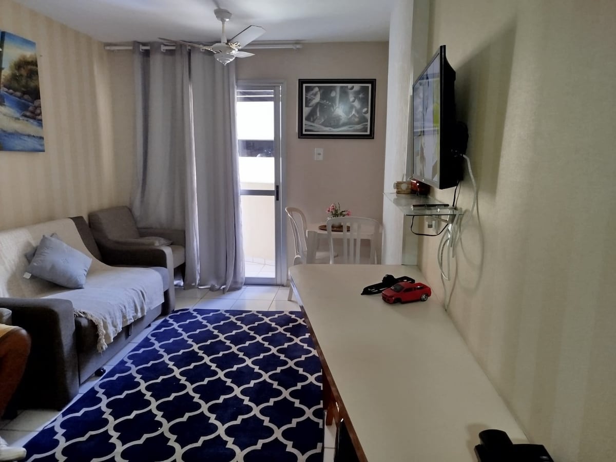 A bright living area features a comfortable sofa paired with light-colored walls and a patterned rug. A glass door allows natural light and leads to a balcony. A small dining table is visible, along with a mounted TV and decorative artwork enhancing the space.