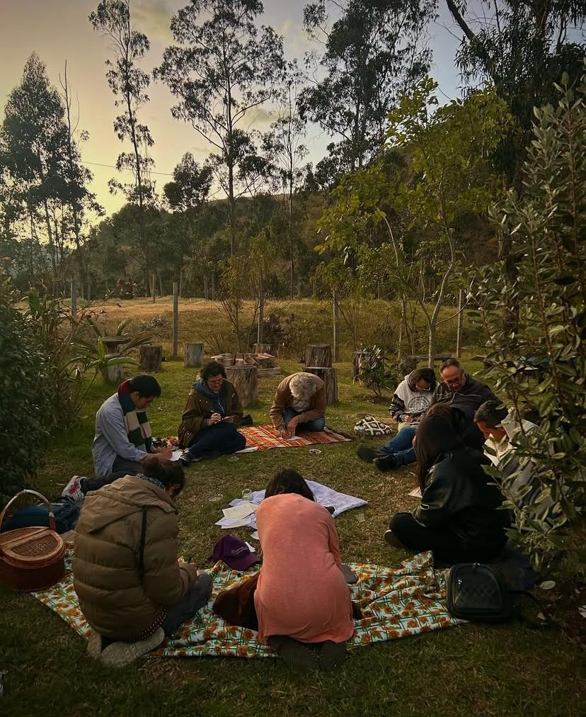 A serene outdoor gathering occurs on a grassy area surrounded by trees. Participants are seated on colorful blankets, engaged in activities. Sunlight filters through the trees, casting gentle shadows. The scene offers a peaceful ambiance, blending nature with communal interaction.