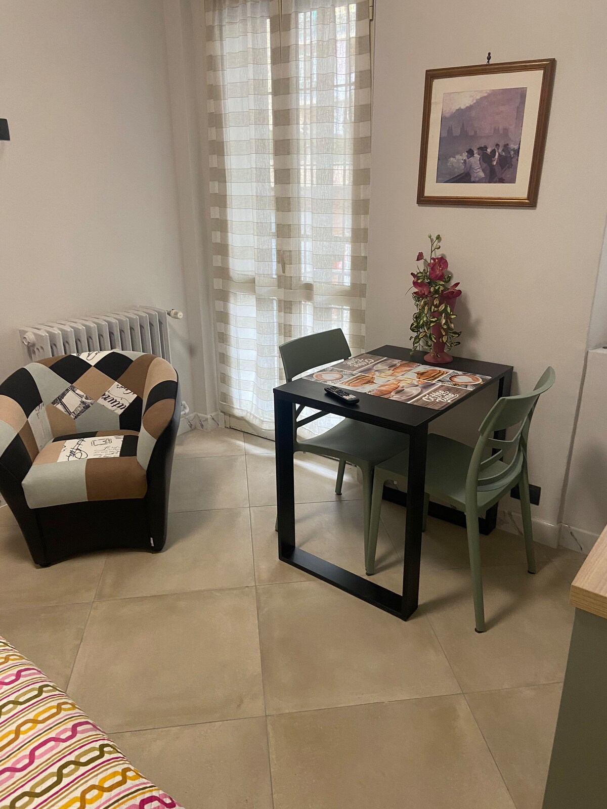 A small dining area features a black table accompanied by two chairs. A patterned armchair offers additional seating. Natural light filters through sheer curtains, illuminating the space. A floral arrangement and framed artwork enhance the room's decor against a neutral backdrop.