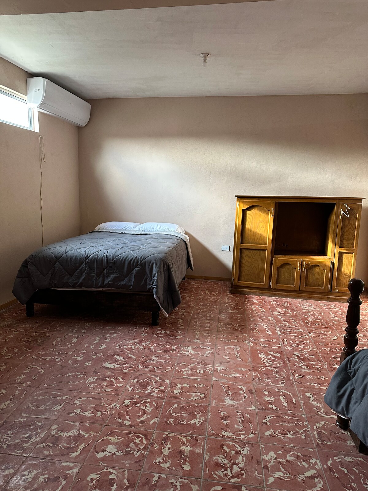 The room features a single bed with a gray quilt and light-colored walls. A wooden cabinet is positioned against one wall, and a window allows natural light to enter. The flooring is a patterned tile, contributing to a cozy ambiance.