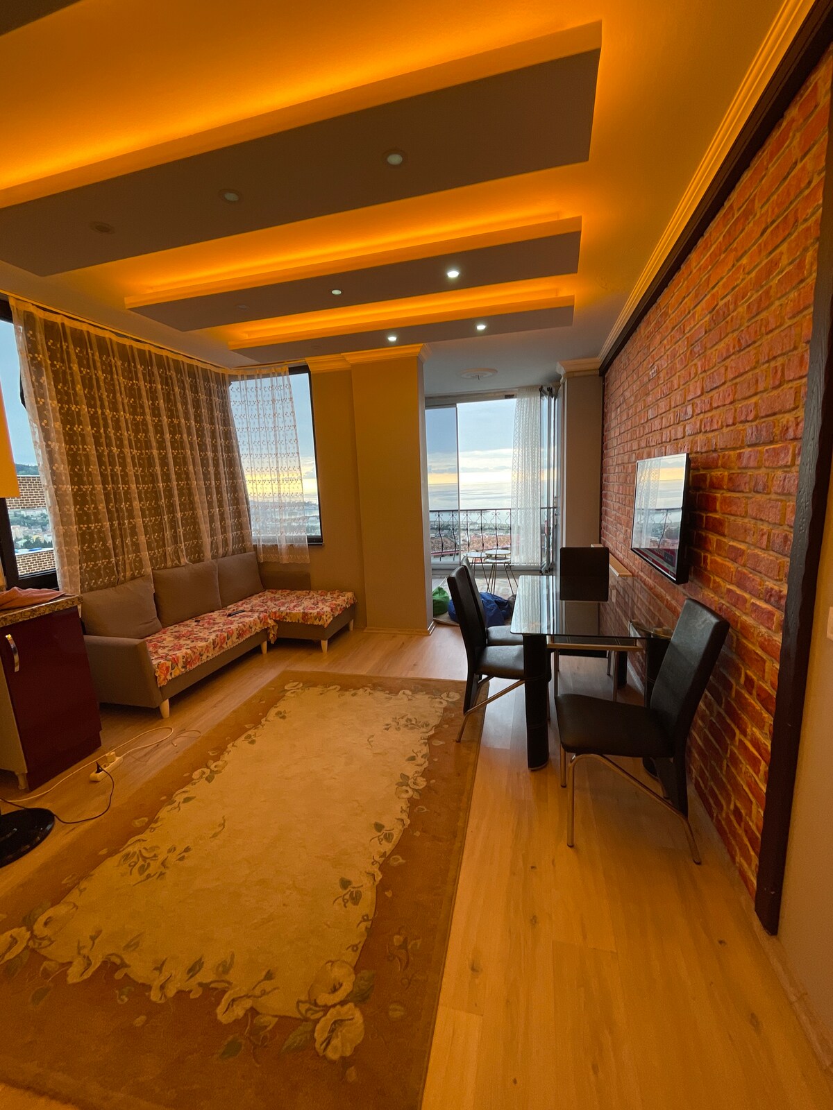 A modern living space features a cozy seating area with a floral-patterned couch and a small dining table for four. Soft lighting highlights the ceiling and creates a warm atmosphere, while large windows offer expansive views of the surroundings.