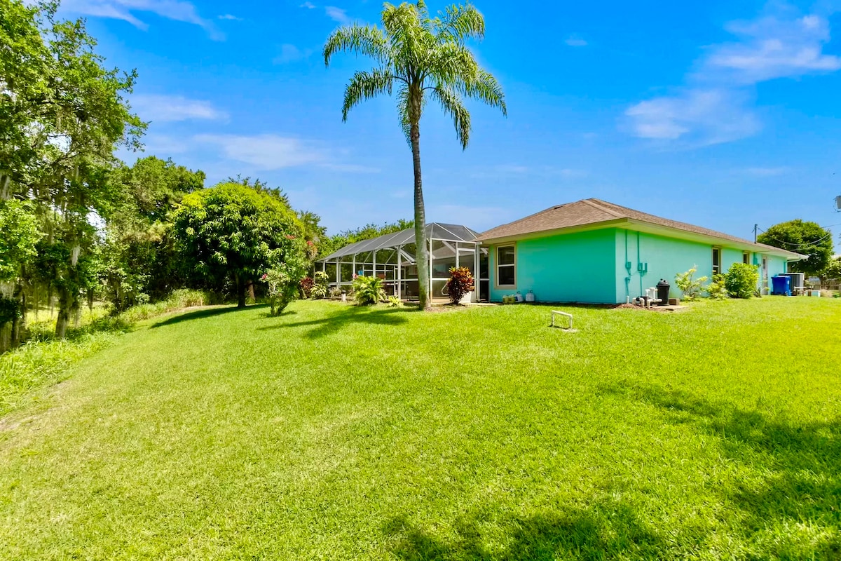 A spacious backyard is visible, featuring a well-maintained green lawn and a tall palm tree. A single-story turquoise house stands in the background, with a screened patio area. Lush greenery surrounds the property, enhancing the outdoor environment.