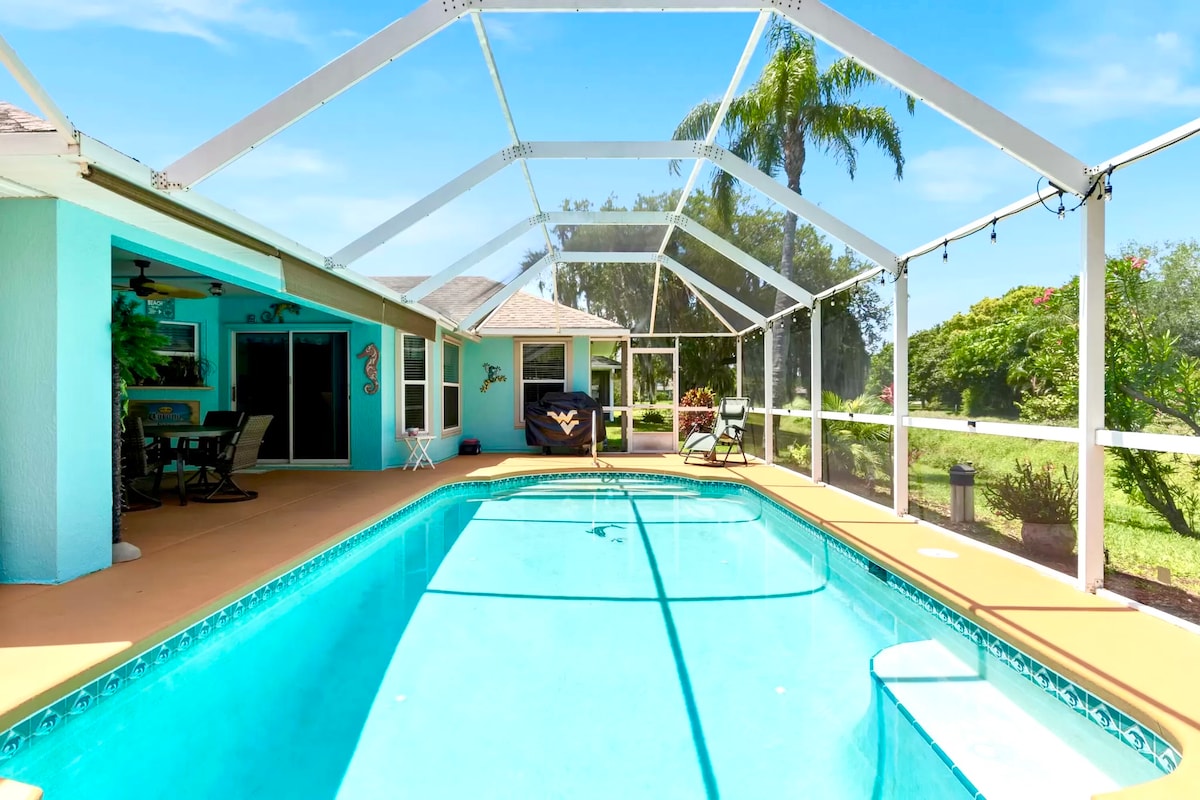 A screened-in pool area is visible, featuring a clear blue pool surrounded by a spacious patio. Light-colored lounge chairs and a small table are positioned near the water. Lush greenery is seen in the background, under a blue sky with a few clouds.