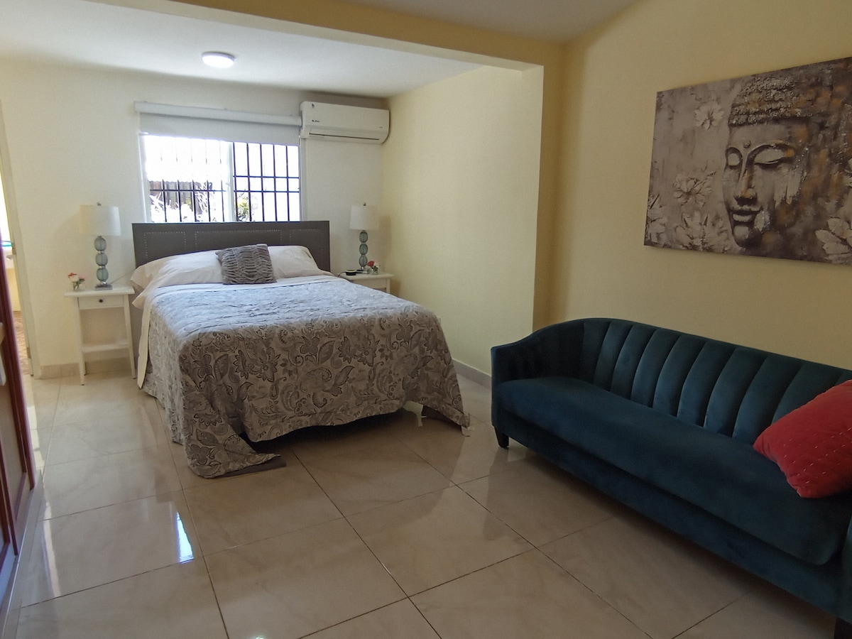 A bright and functional room features a spacious bed with a decorative coverlet. A cozy couch accompanies the seating area, while natural light enters through a window. The walls are painted in soft tones, contributing to a serene atmosphere.
