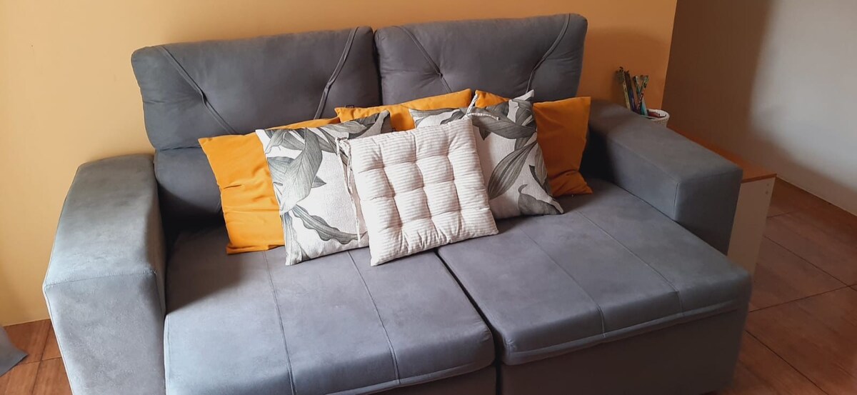 A comfortable gray sofa is positioned against a warm orange wall. It features multiple cushions in soft neutral tones and a few decorative elements. The sofa appears inviting, with ample seating space and a modern design.