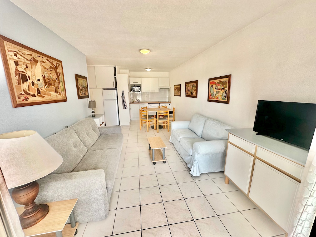A spacious living area features comfortable seating arrangements with two sofas and a wooden coffee table. The kitchen is visible, equipped with modern appliances. Decorative artwork adorns the walls, adding a touch of character, while natural light fills the space through large windows.