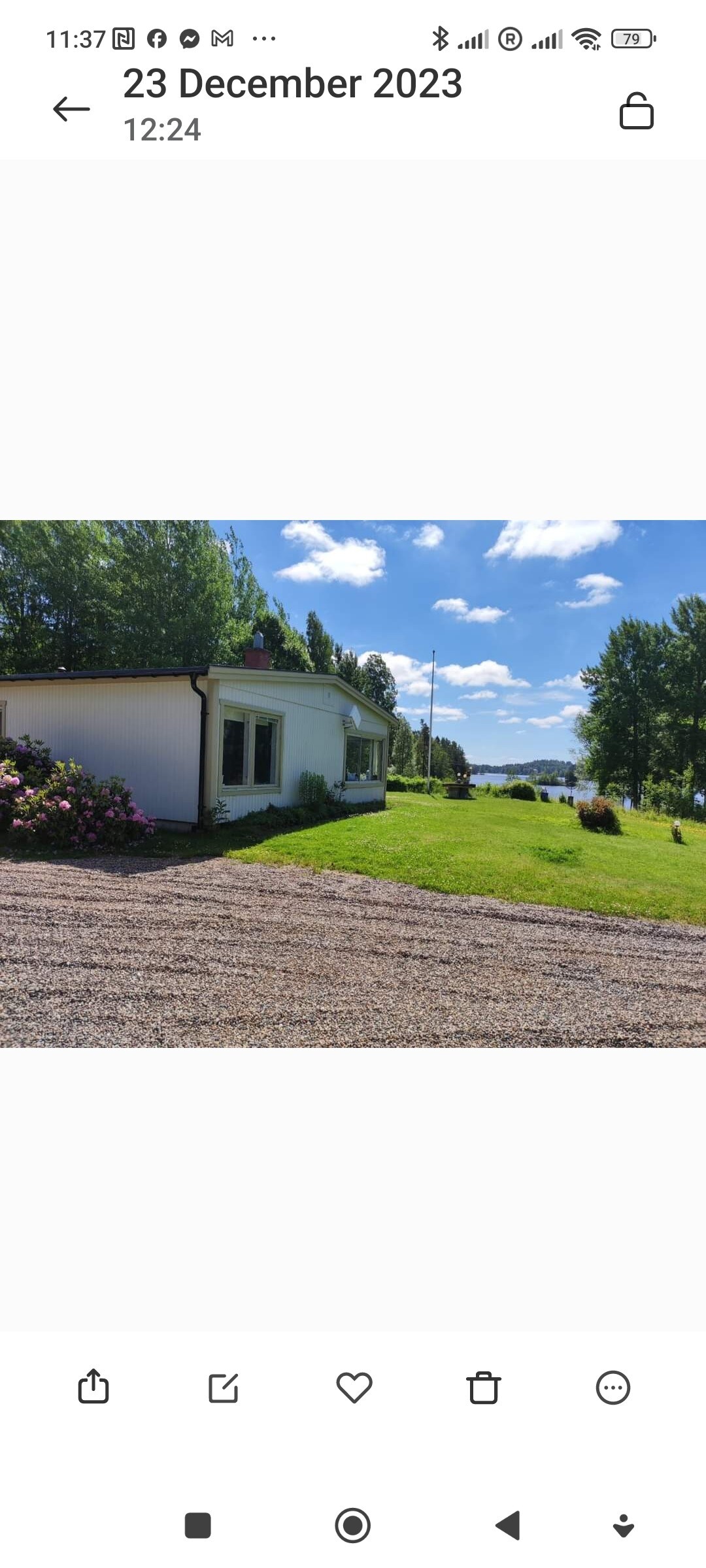The exterior of the property is presented, featuring a white building framed by green grass. A well-maintained garden with colorful flowers is visible in the foreground. The background includes a serene view of a lake and a clear blue sky with scattered clouds.