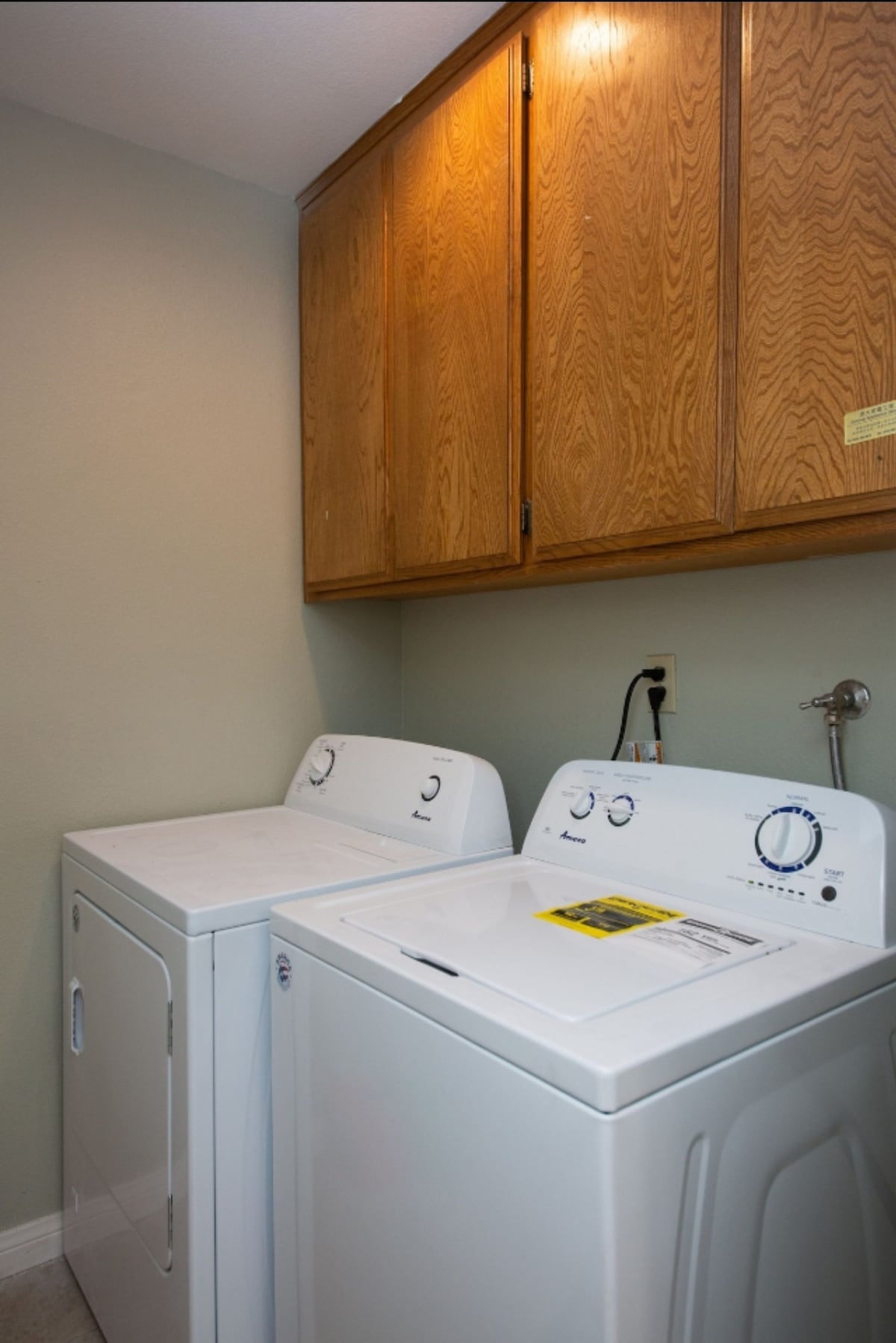 A laundry area is equipped with a white washing machine and dryer. Above, wooden cabinets provide storage space. The wall is painted a soft green, contributing to a clean and functional appearance.