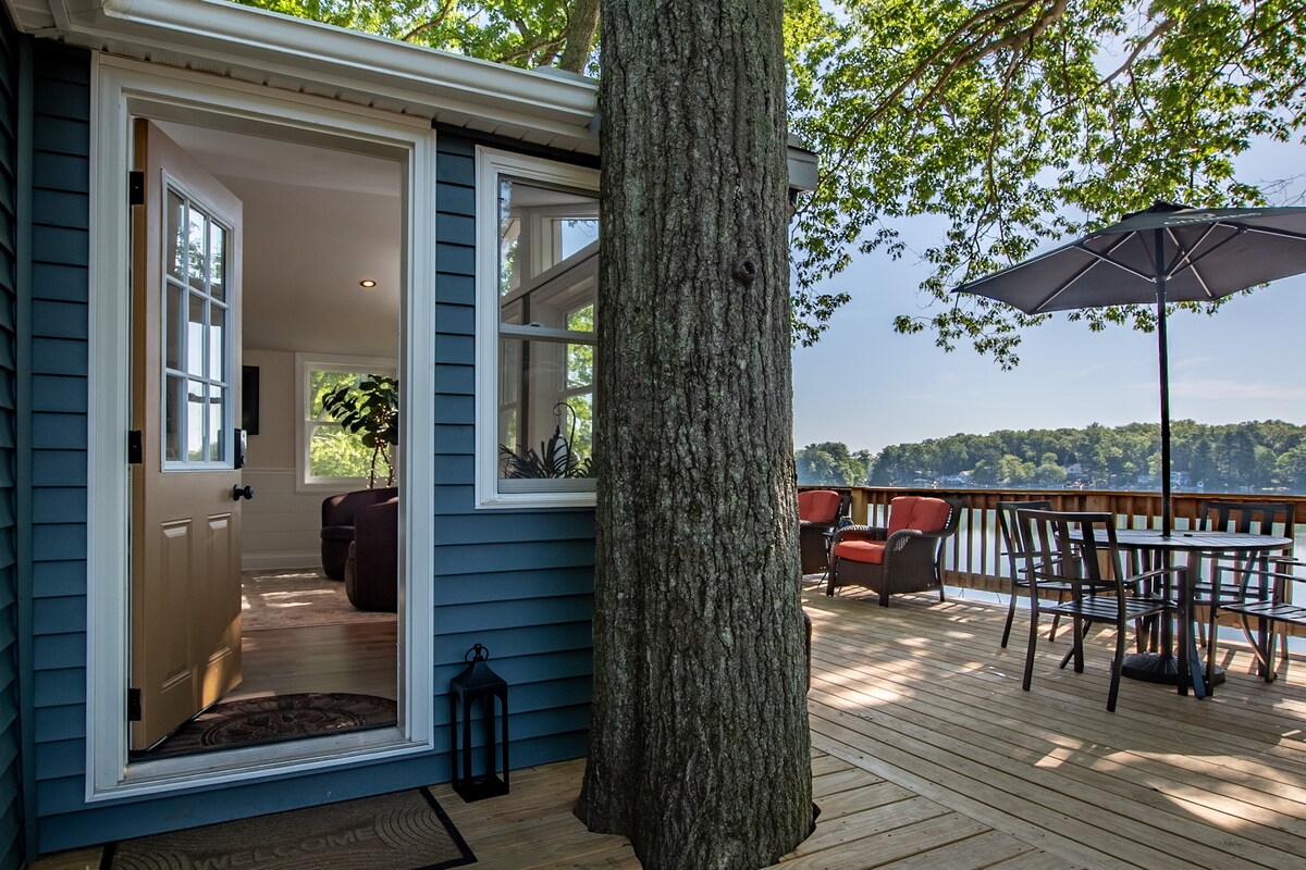 The entrance of the cottage is framed by a large tree, leading to a spacious deck overlooking the lake. Outdoor seating is arranged around a table, with a shaded area provided by an umbrella. Bright natural light enhances the cozy atmosphere.