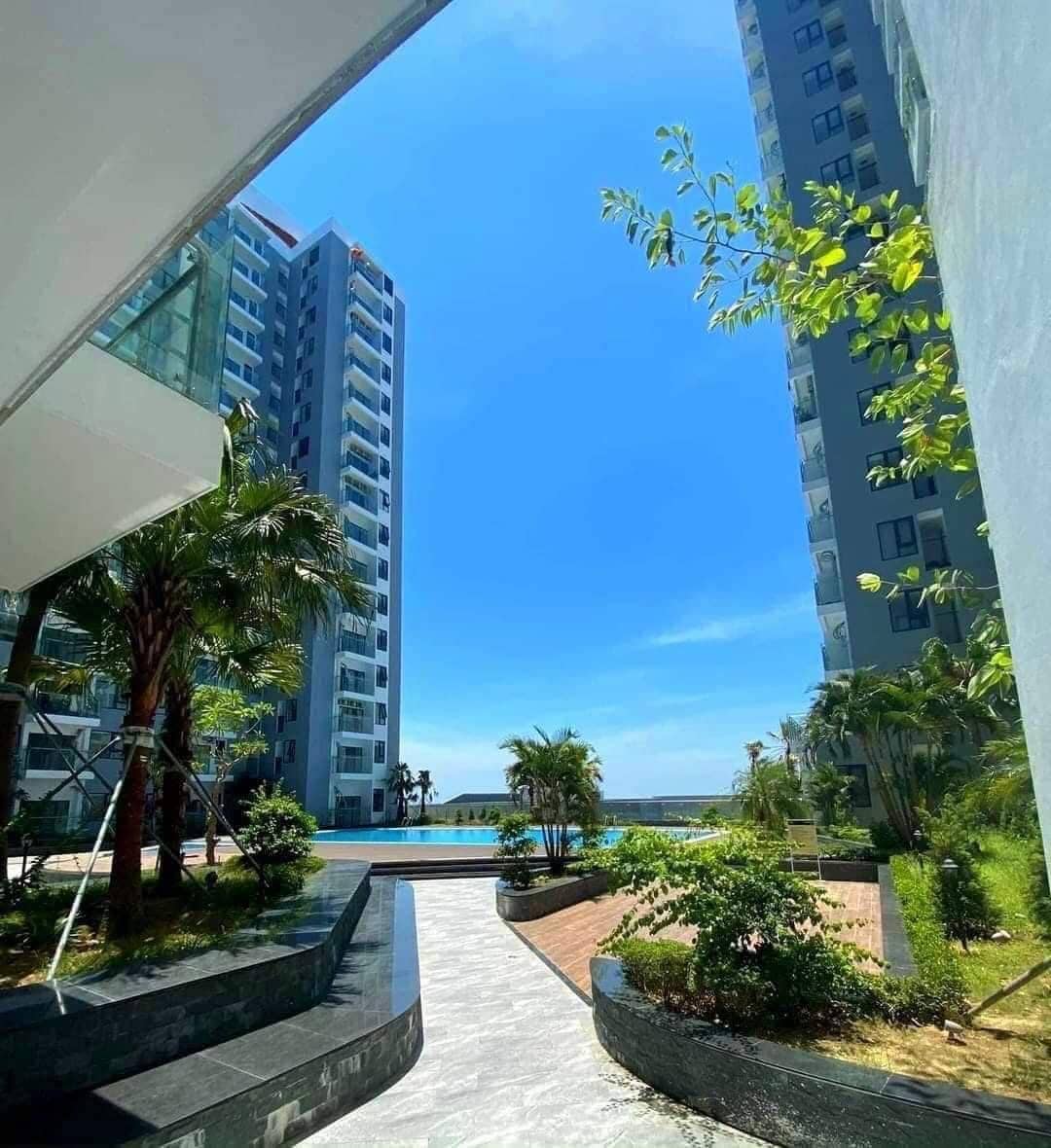 A modern pathway leads between two buildings, flanked by lush greenery and tropical plants. The clear blue sky above enhances the bright atmosphere, and a swimming pool is glimpsed in the distance, creating a sense of openness and tranquility.