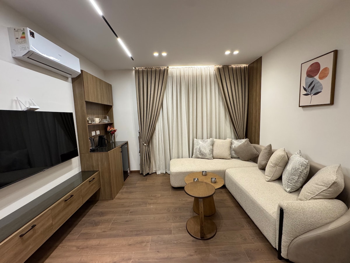 A modern living area is presented with a spacious sectional sofa adorned with various textured cushions. A wooden coffee table sits centrally, complemented by a flat-screen TV mounted on the wall. Soft natural light filters through floor-length curtains, enhancing the inviting ambiance.