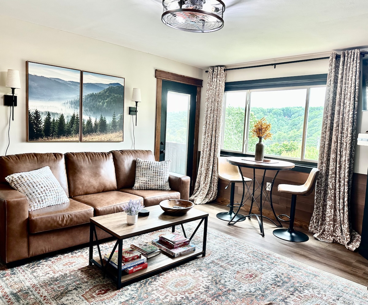 A cozy living area contains a brown leather sofa adorned with patterned pillows and a coffee table featuring books and decorative items. Large windows provide sweeping mountain views, while elegant curtains frame the scene. A dining table for two is positioned nearby, enhancing the inviting space.