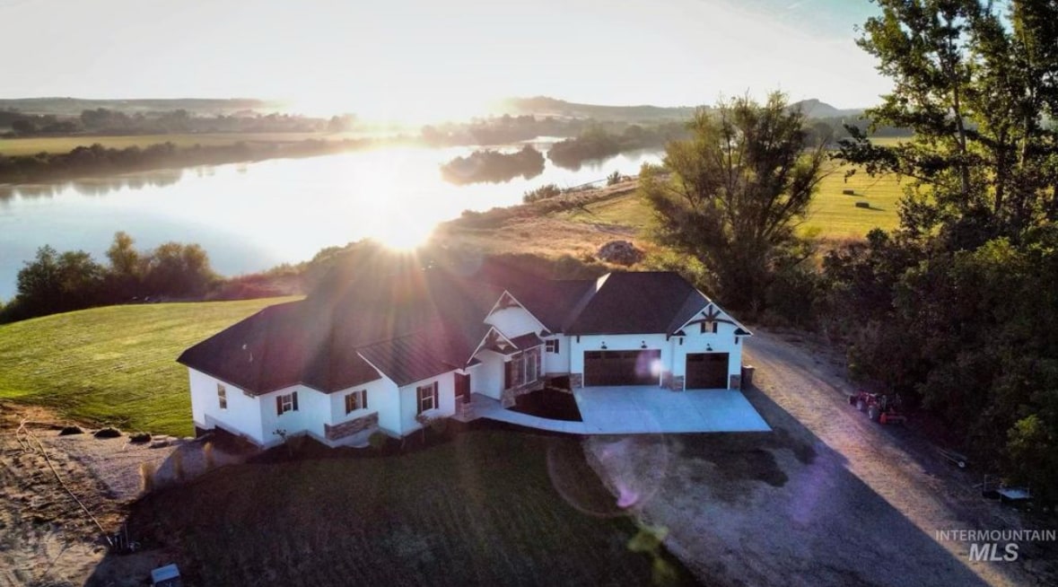Aerial view of a spacious home situated near a river, with the morning sun reflecting off the water. Surrounding greenery and open fields create a serene setting. The house features a combination of a dark roof and light-colored siding, with a driveway leading up to a garage.
