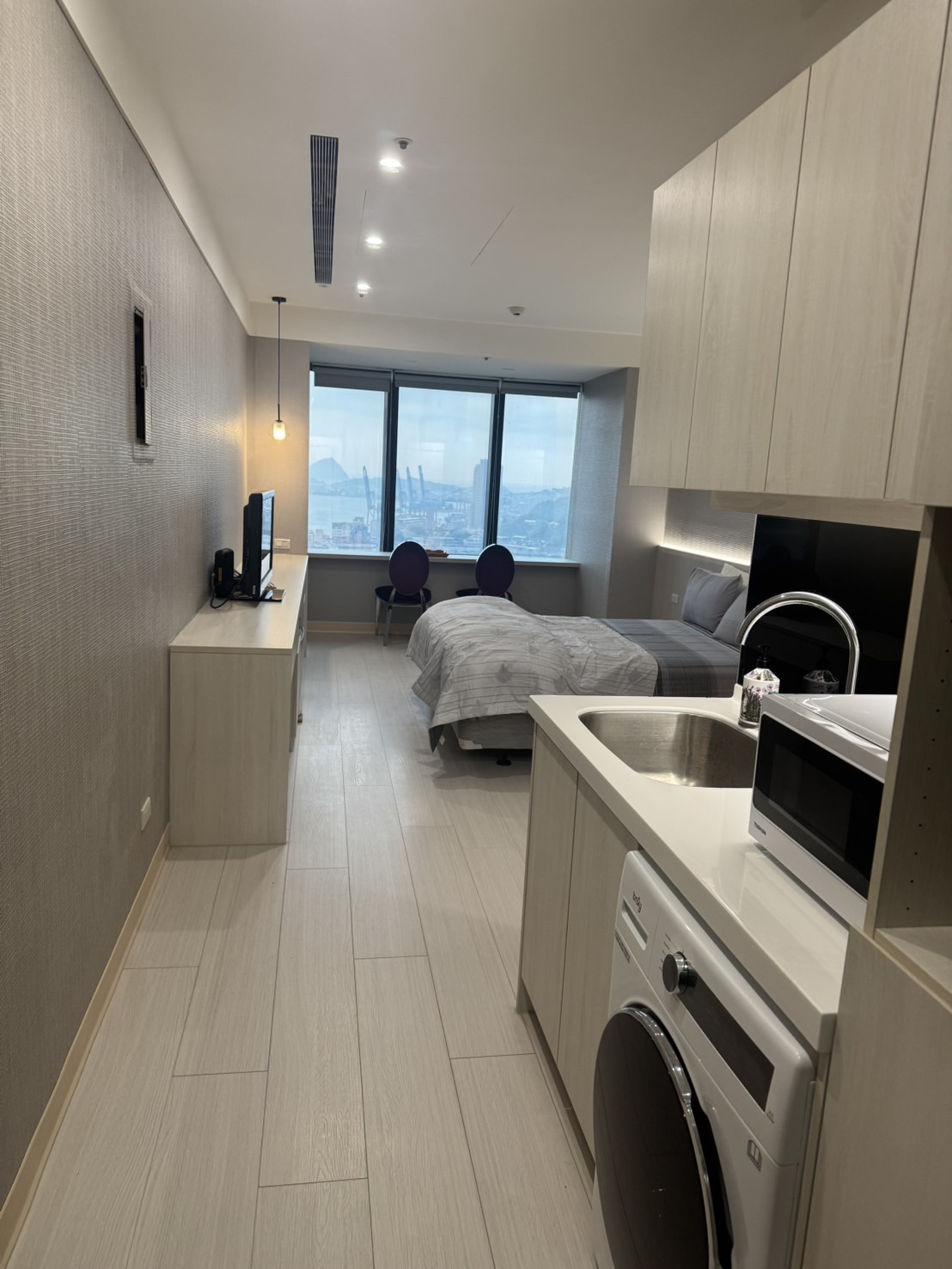 A modern studio apartment is displayed, featuring a compact kitchenette with a sink, microwave, and washing machine. Two beds are neatly arranged at the far end, while a small table with two chairs is positioned next to a large window showcasing a city view.