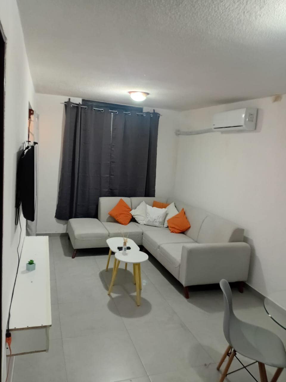 A well-lit living area features a light-colored sectional sofa with orange accent pillows. A small white table is positioned in front. A wall-mounted TV is visible, and a dining table with chairs complements the space. Dark curtains frame the window, enhancing the room's comfort.