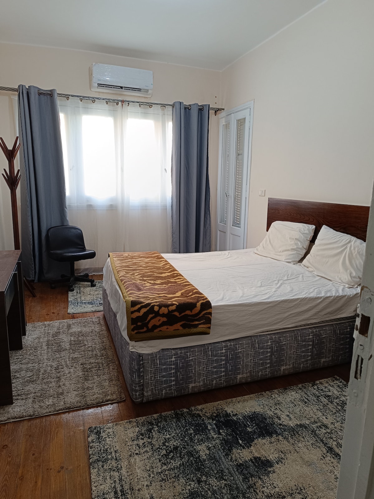 A well-lit bedroom features a double bed with a brown and gold throw blanket. A desk and a chair are positioned nearby, complemented by patterned rugs on the wooden floor. Large windows framed by light curtains allow natural light to illuminate the space.