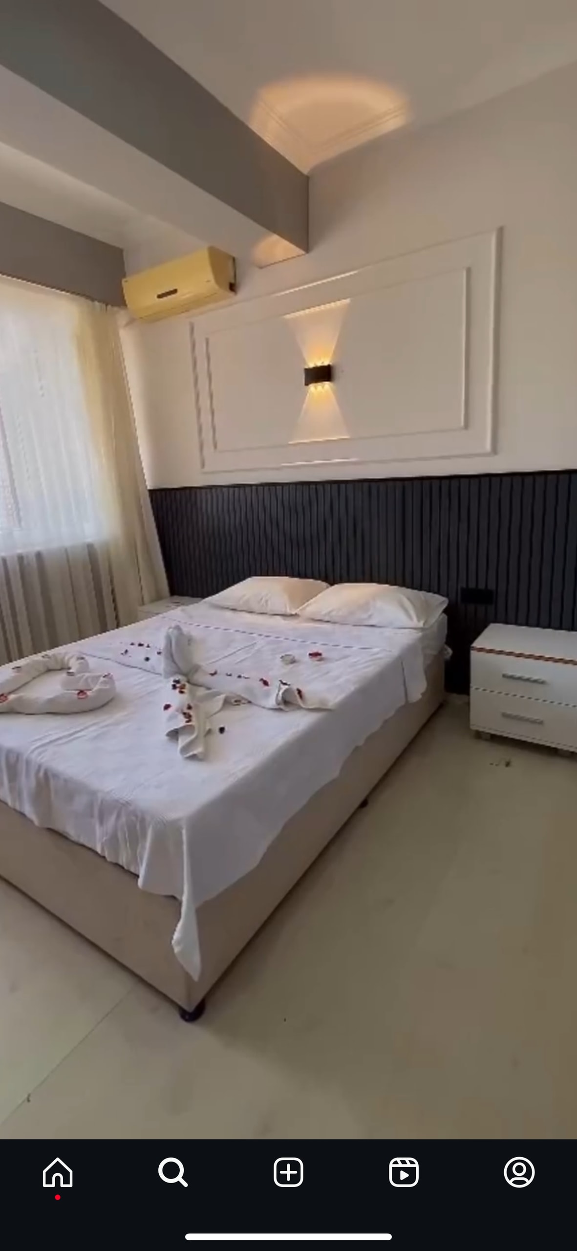 A bright sleeping area features a spacious bed dressed in light linens, with decorative rose petals scattered across the cover. A bedside table stands next to the bed, and gentle light filters through sheer curtains, enhancing the calm ambiance of the room.