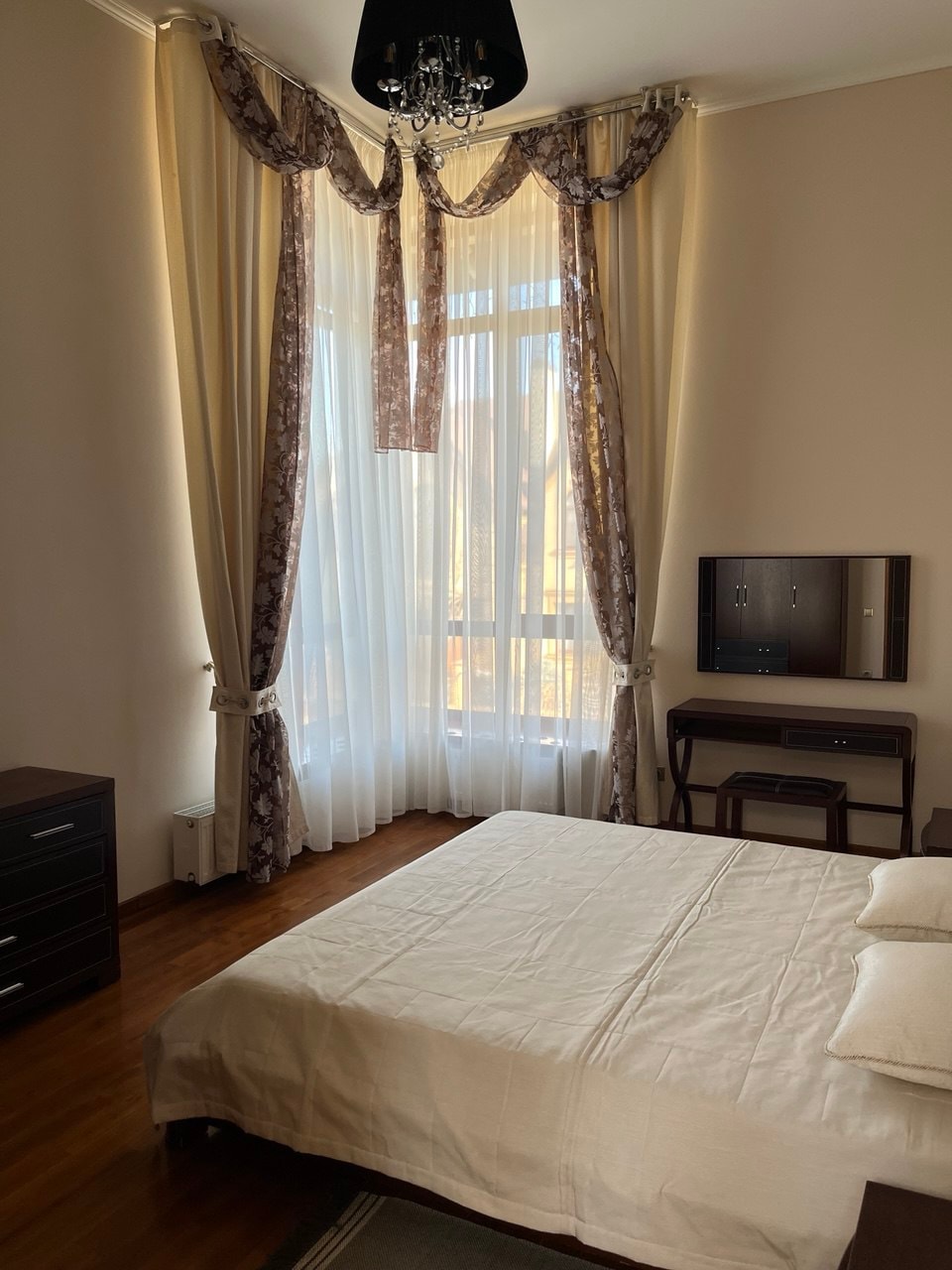 A bright sleeping area features a spacious bed dressed in neutral linens. Tall windows are adorned with sheer curtains that allow natural light to filter in, complemented by decorative drapes. A dark wooden dresser and a small desk are positioned in the corner.